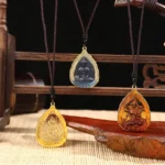 Multiple handcrafted Buddhist Liuli crystal pendants in vibrant colors displayed on wooden rack with bamboo mat, showing orange, blue, and green Buddha designs with gold metal frames