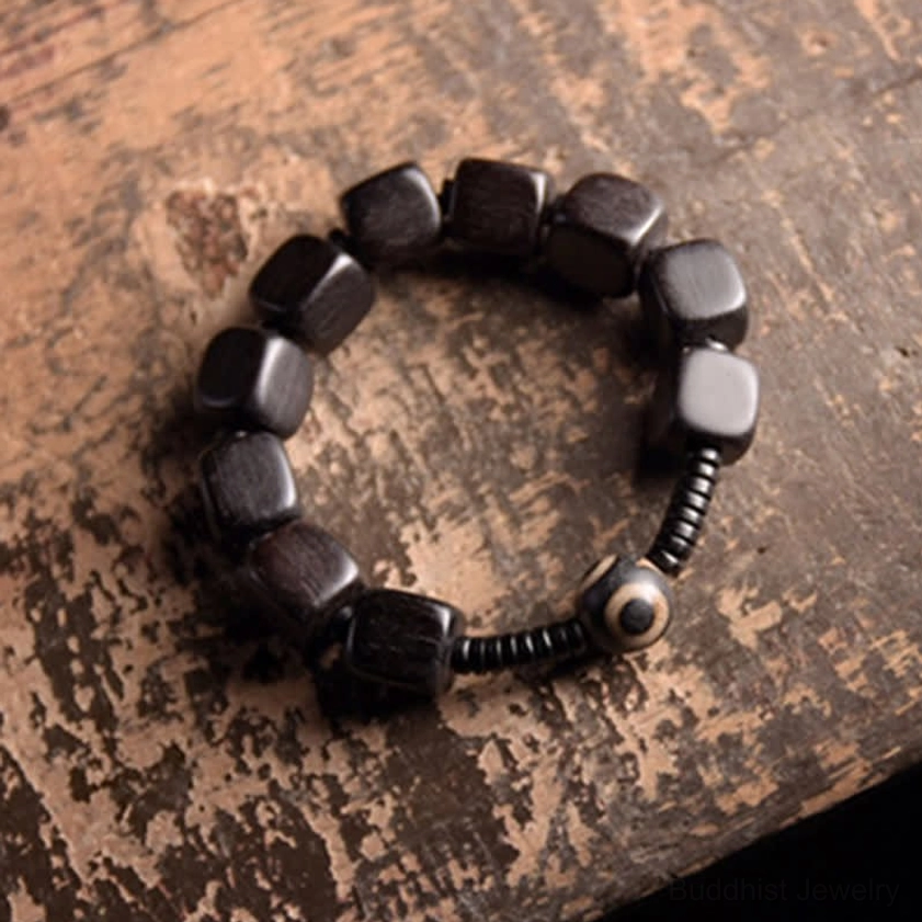 Buddhist ebony dzi bead bracelet featuring dark cubic beads and sacred eye charm on rustic wooden backdrop