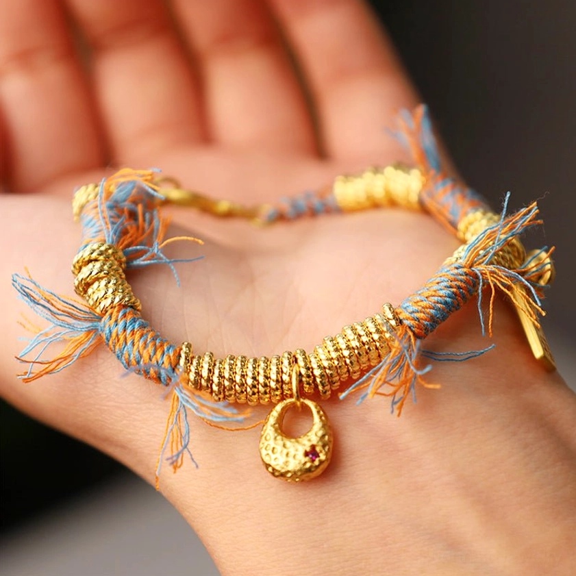 Buddhist braided bracelet laid on human palm displaying complete design with round gold charm, frayed tassel details, and natural string construction