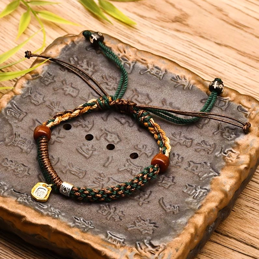 Green braided Buddhist protection bracelet with wooden beads and gold charm, displayed on natural bamboo background