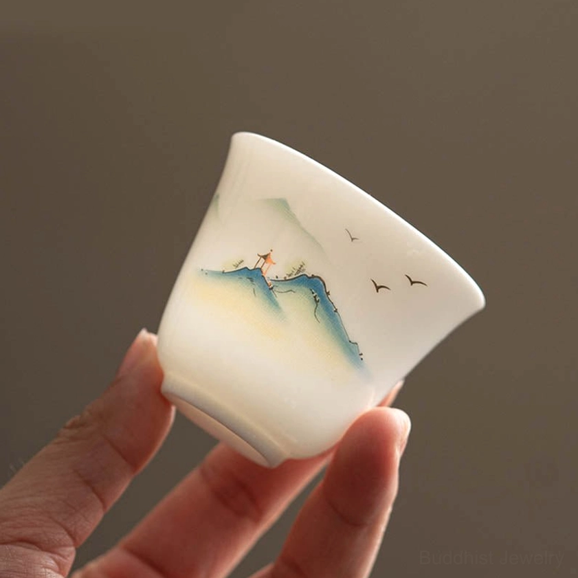 Hand-held Buddhist ceramic tea cup with clear hand-painted mountain and bird landscape artwork against neutral background
