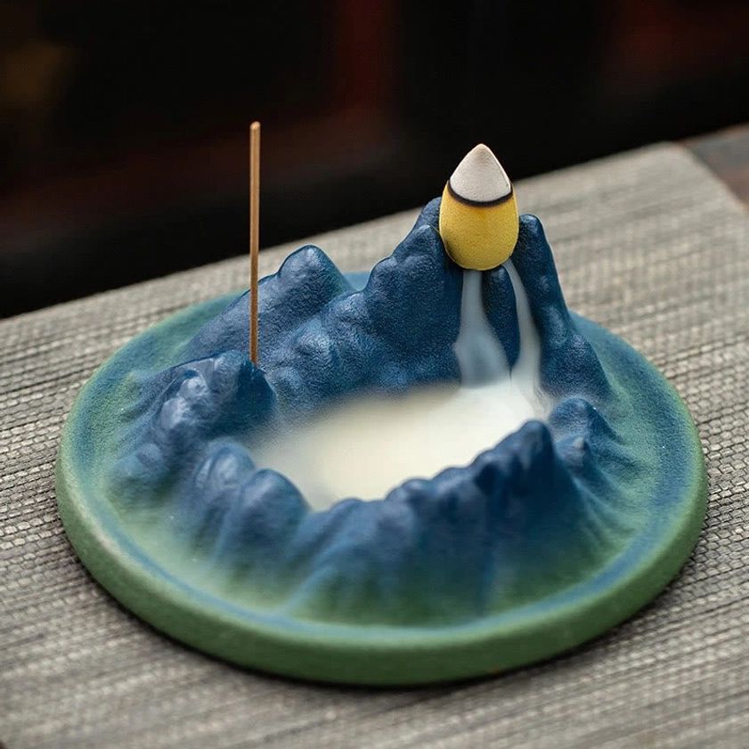 Ceramic Buddhist incense holder with active backflow smoke cascading down mountain landscape, displayed on linen placemat