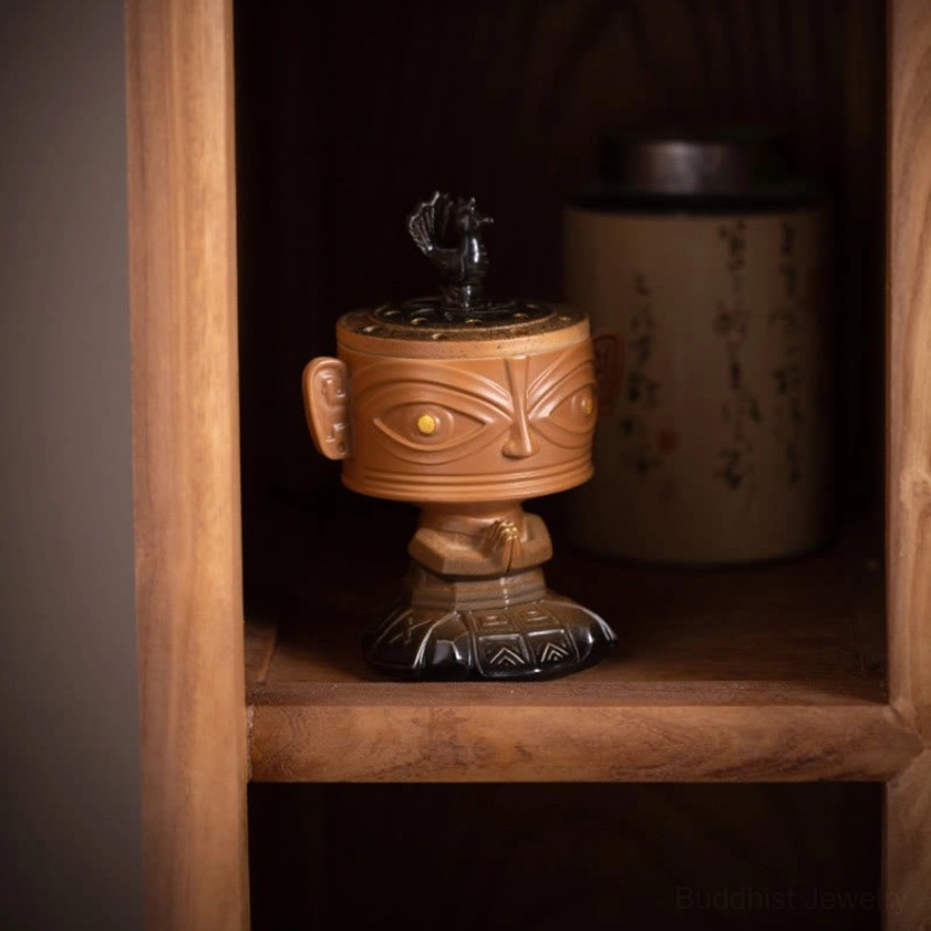 Buddhist ceramic incense burner with bronze face design displayed on wooden shelf with tea accessories