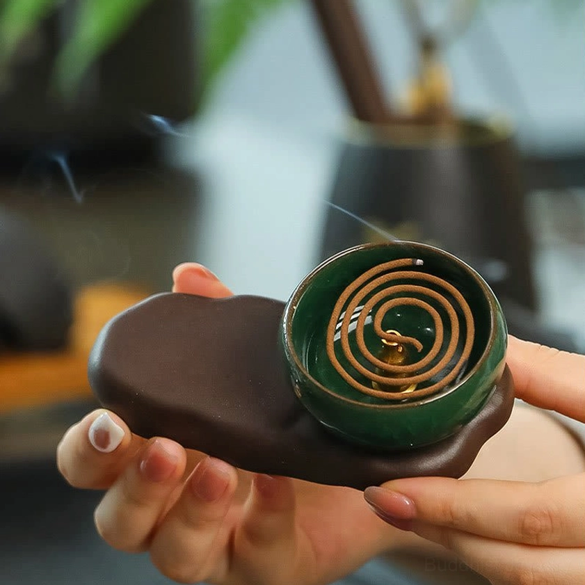 Ceramic incense holder held in hands displaying spiral coiled incense inside green glazed bowl with stone-shaped brown ceramic base