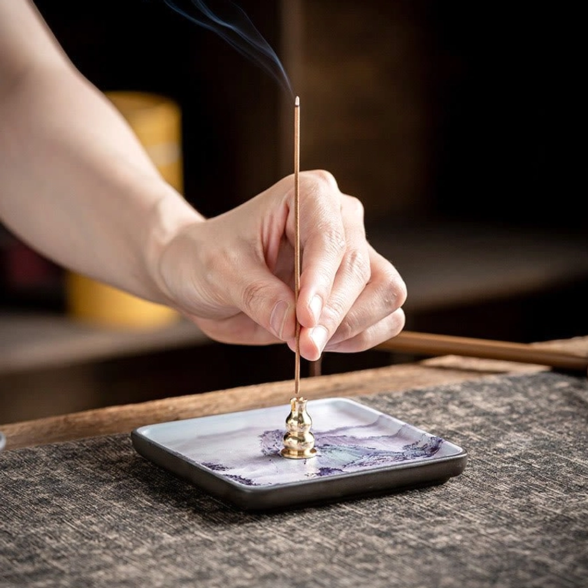 Hand placing stick incense onto brass holder in handcrafted ceramic Buddhist incense burner tray