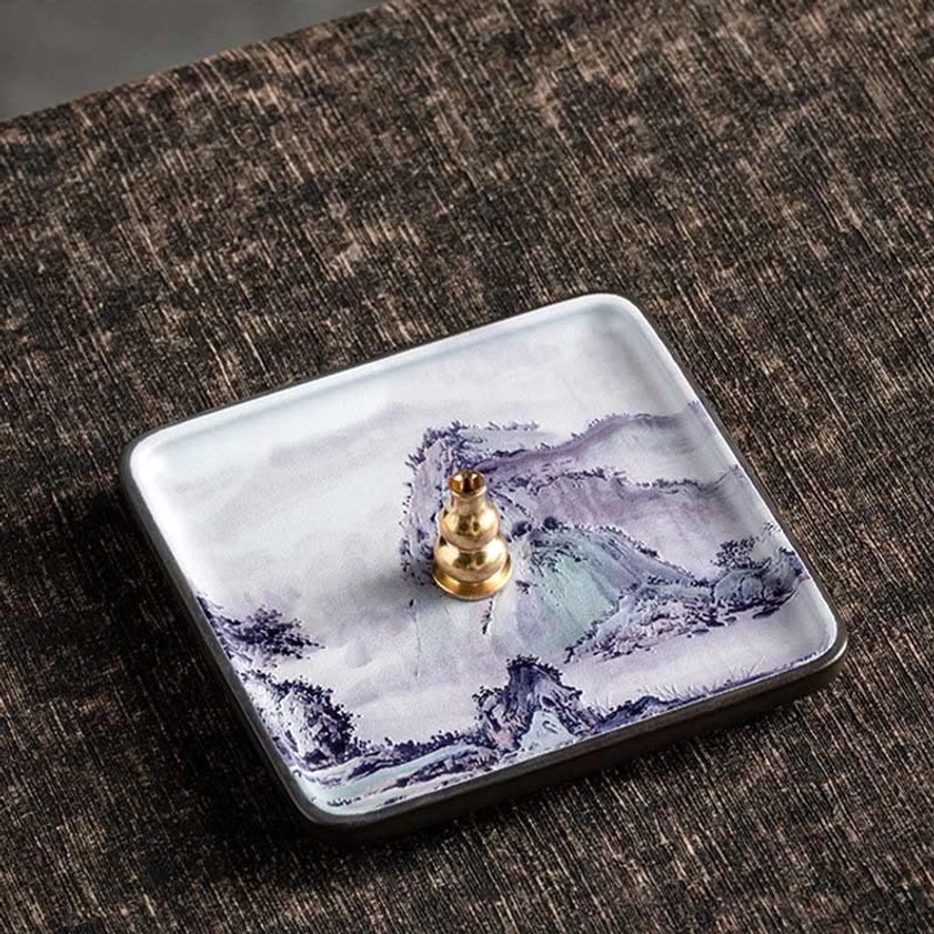 Square ceramic incense burner tray with Buddhist landscape motif on linen fabric tabletop with brass holder detail