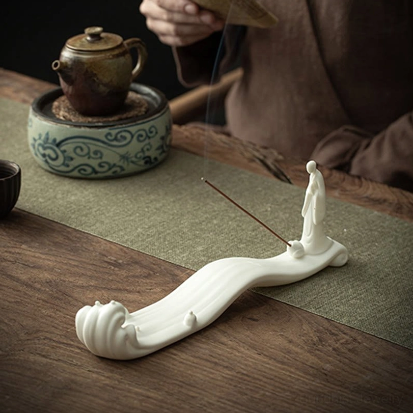 White ceramic Buddhist incense holder on wooden tea table with ceramic tea ware and burning incense