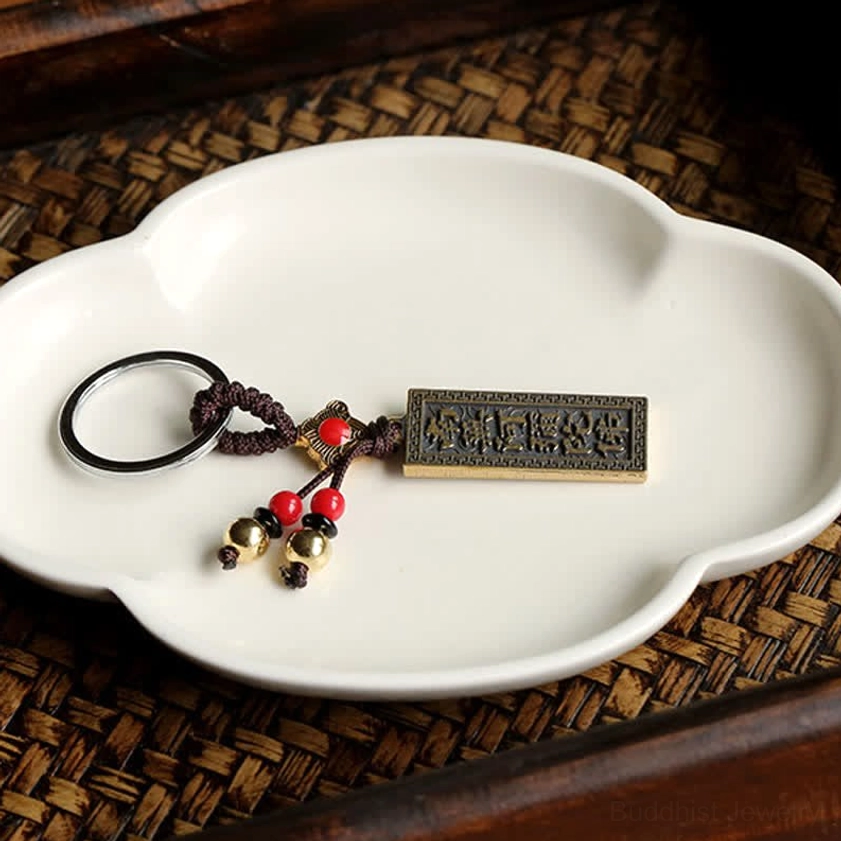 Top-down flat lay of Buddhist blessing keychain with red bead tassel and brass bells on woven mat with ceramic plate