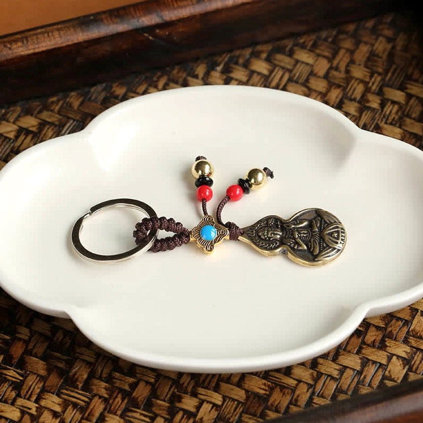 Top-down overhead view of handmade Buddha blessing keychain on decorative ceramic flower plate, showing mixed colored beads in red, blue, and gold with flat bronze gourd charm