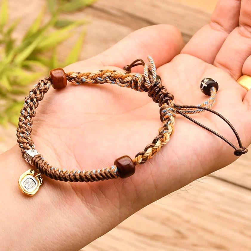 Khaki-brown Buddhist protection bracelet in hand with bamboo foliage, showing adjustable sliding knots and charm pendant