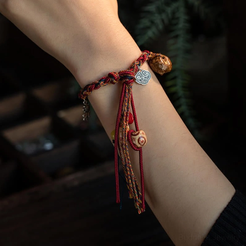 Buddhist protection bracelet worn on wrist showing red braided rope, silver Ruyi charm, and tassel detail, close-up side view against green foliage background