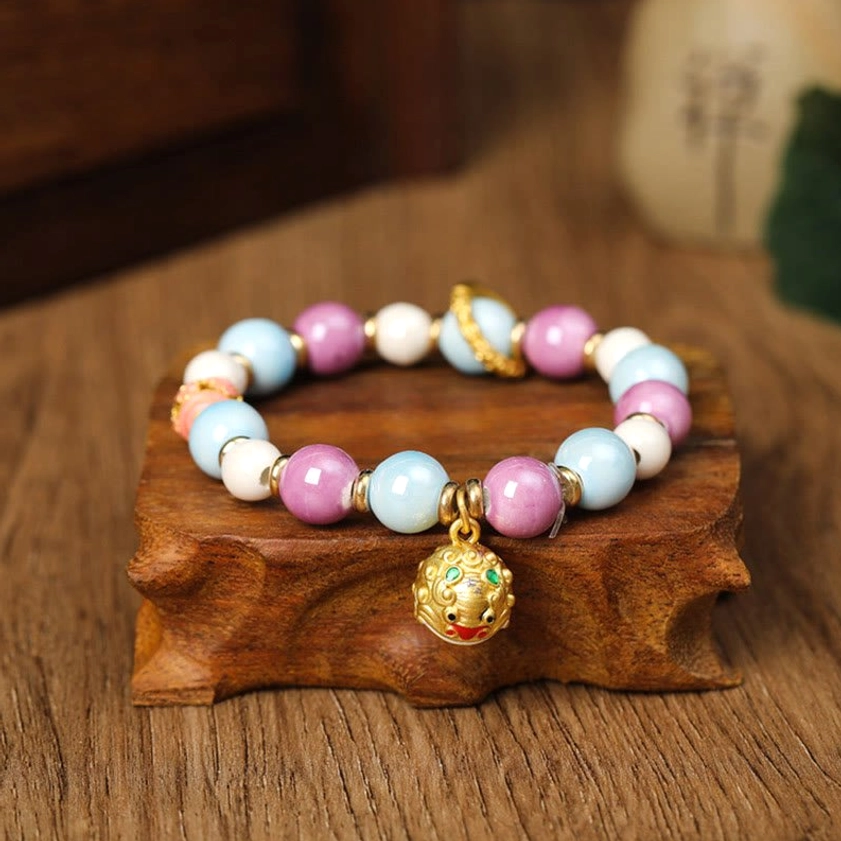 Buddhist prosperity bracelet with lion head gold charm pendant and pastel beads displayed on wooden table with decorative stand