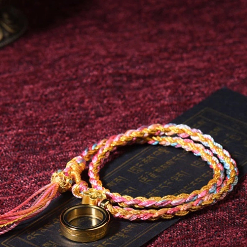 Single wrap Buddhist lucky braided string bracelet with pink-gold threads, metal ring charm and flower decoration on dark red cloth