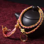 Handmade Buddhist lucky braided string bracelet with multicolor threads, metal flower charm and tassel - main product view on dark red cloth