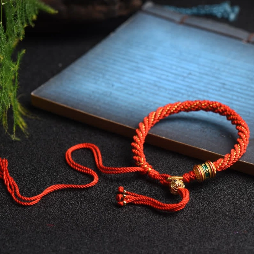 Red Buddhist string bracelet flat lay on vintage paper with green plants, woven rope and gold metal accessories