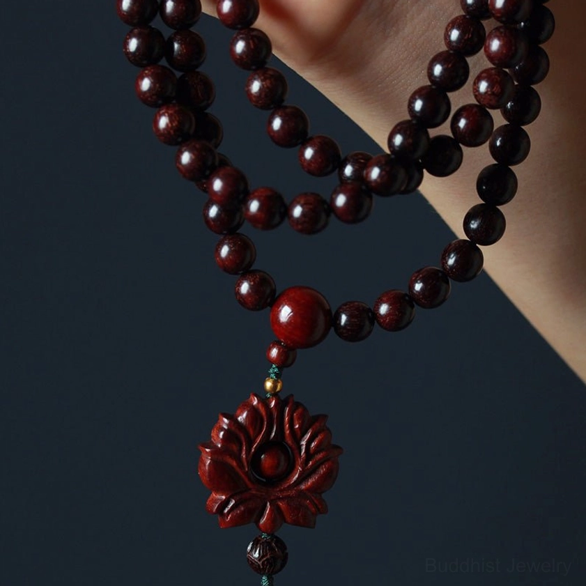 Hand-held side view of sandalwood prayer mala showing natural wood grain texture lotus pendant with hanging tassel and red accent bead
