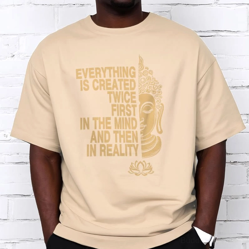 Man wearing light tan Buddhist graphic tee with black pants and gold print against white brick wall