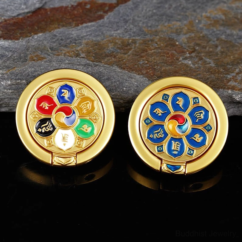 Two Buddhist mantra phone ring holders in different enamel patterns displayed flat on stone surface, gold-plated metal construction