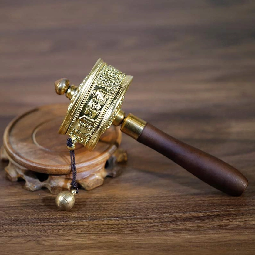 Handheld Buddhist prayer wheel with engraved sutra patterns, gilt metal construction, dark wood handle on wooden display stand - three quarter view