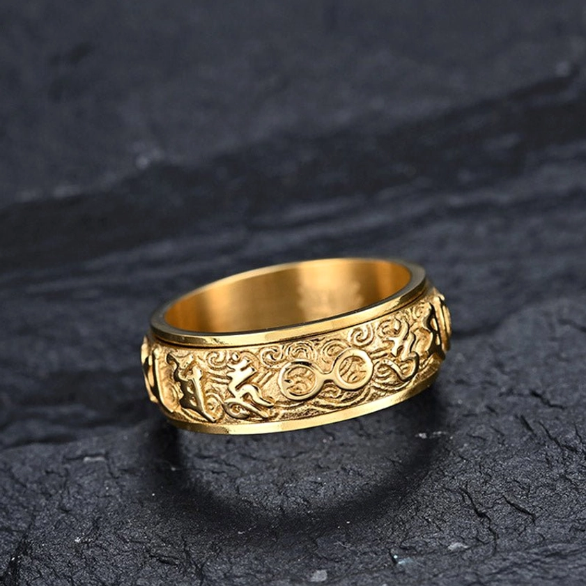 Gold-finished Buddhist mantra ring with intricate scroll and Sanskrit engravings, front view on dark slate background showing craftsmanship