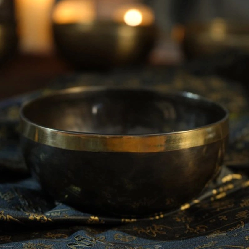 Close-up Buddhist singing bowl with worn dark metal surface and bright polished gold rim on candlelit patterned cloth backdrop