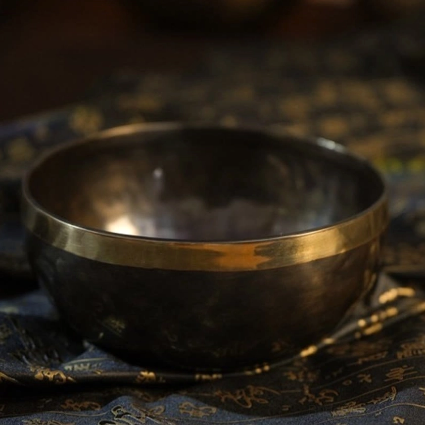 Low angle three-quarter view of handmade Buddhist singing bowl with matte dark bronze body and polished gold rim on patterned textile