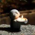 Buddhist monk resin candle holder with lit tealight on outdoor stone surface - front three-quarter view showing grey matte finish