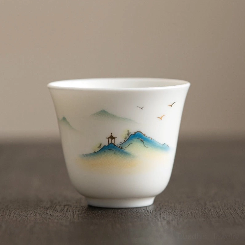 Buddhist-inspired landscape porcelain tea cup with hand-painted mountains, pavilion, and flying birds, shown on dark wooden table surface