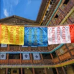 Buddhist prayer flags set hanging outdoors in traditional Tibetan architectural setting, showing multi-color satin flags with printed mantras and Windhorse motifs