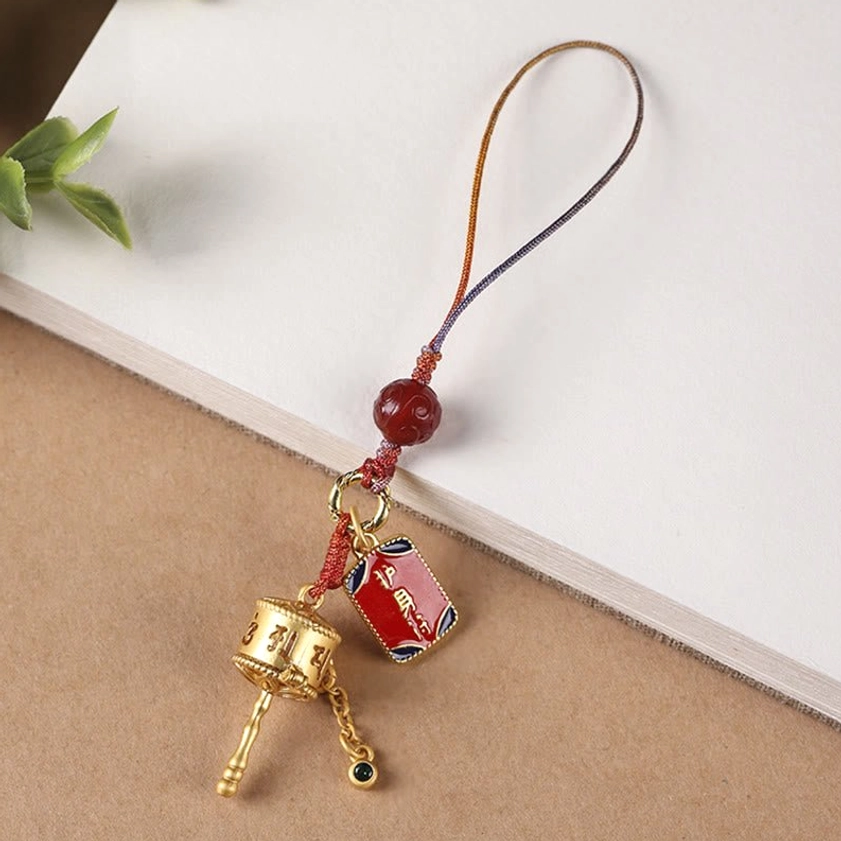 Buddhist prayer wheel phone charm with gradient braided strap and gold-plated ornament on book desktop