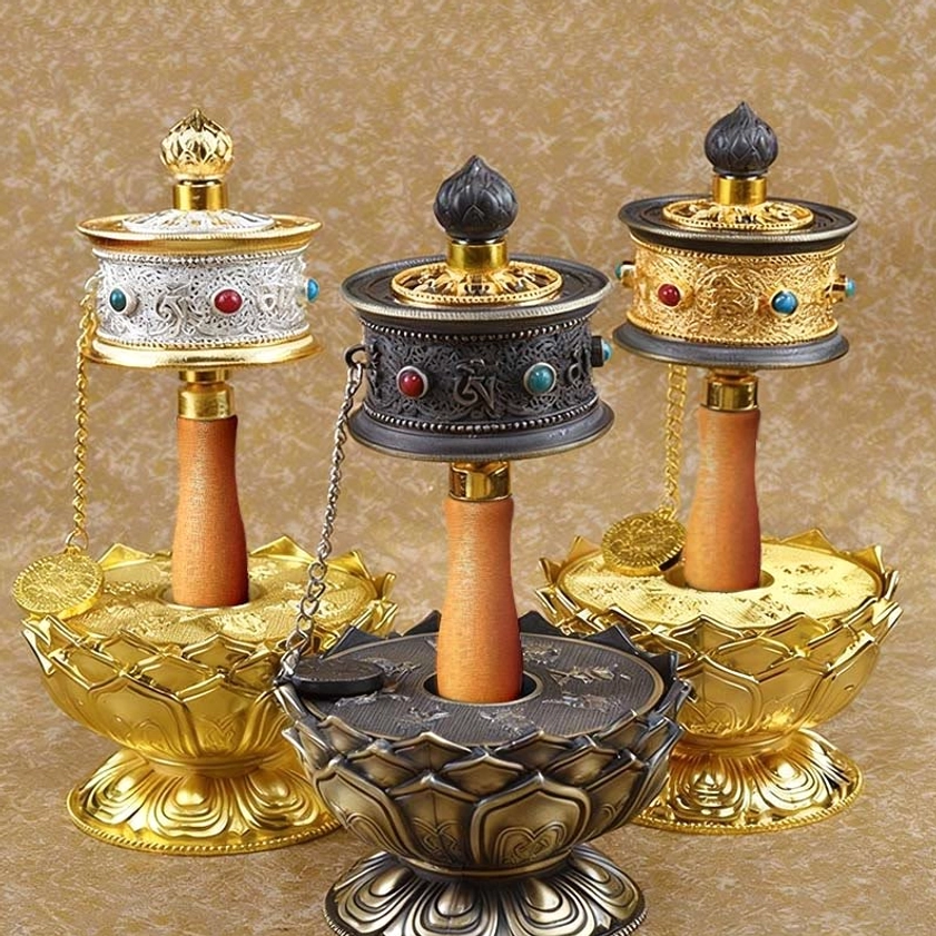 Three Buddhist prayer wheels in varied styles featuring silver, bronze, and gold finishes displayed with decorative lotus base stands