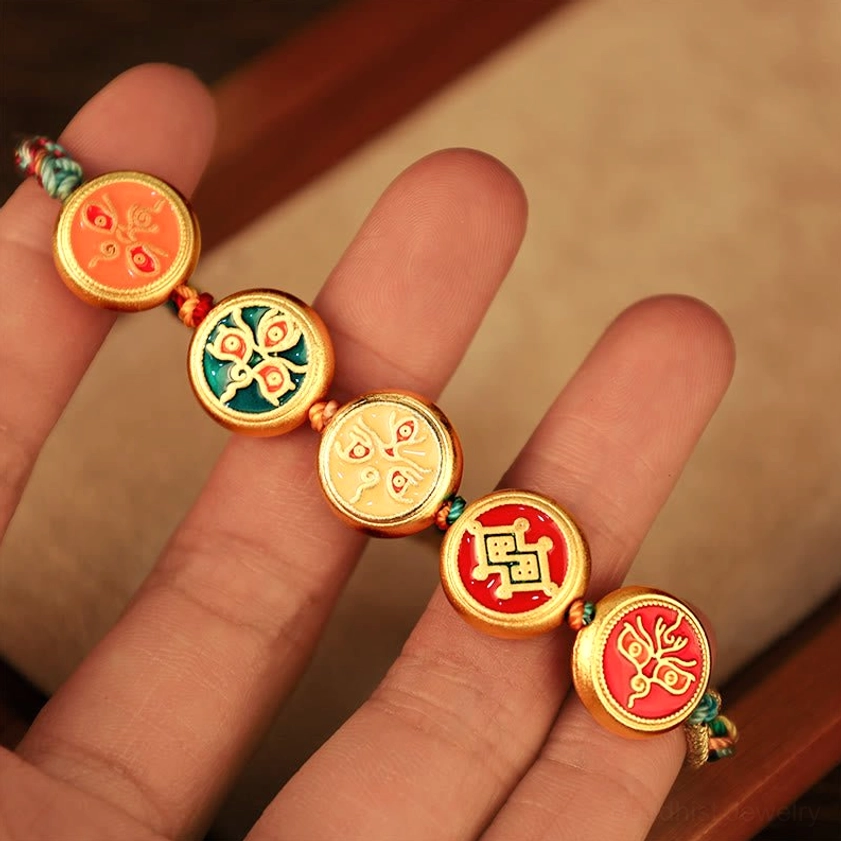 Buddhist wealth deity enamel charms in close-up held on fingertips, showing five gold-plated charms with traditional iconography