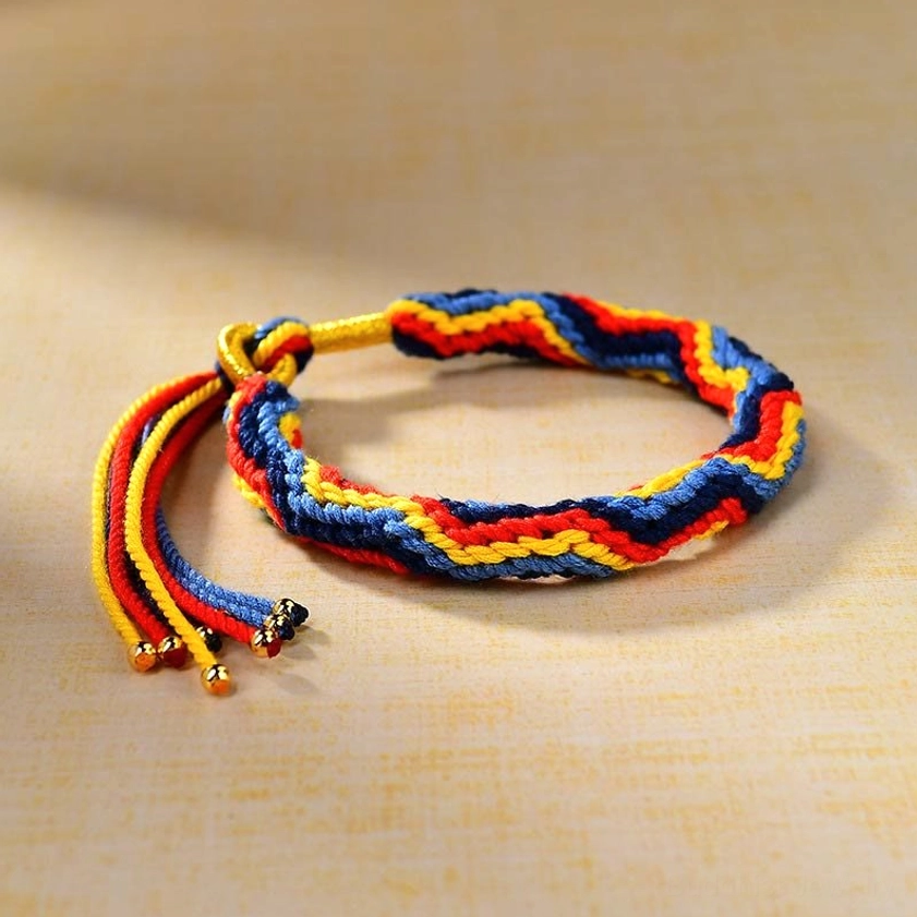 Flat lay view of hand-braided lucky Buddhist bracelet displaying striped colorful weave pattern with adjustable knot closure and gold beaded tassels on light beige background