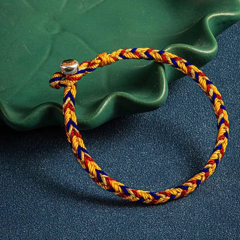 Handwoven multicolored Buddhist protection bracelet displayed on green wavy tray, showing braided rope texture and metal charm