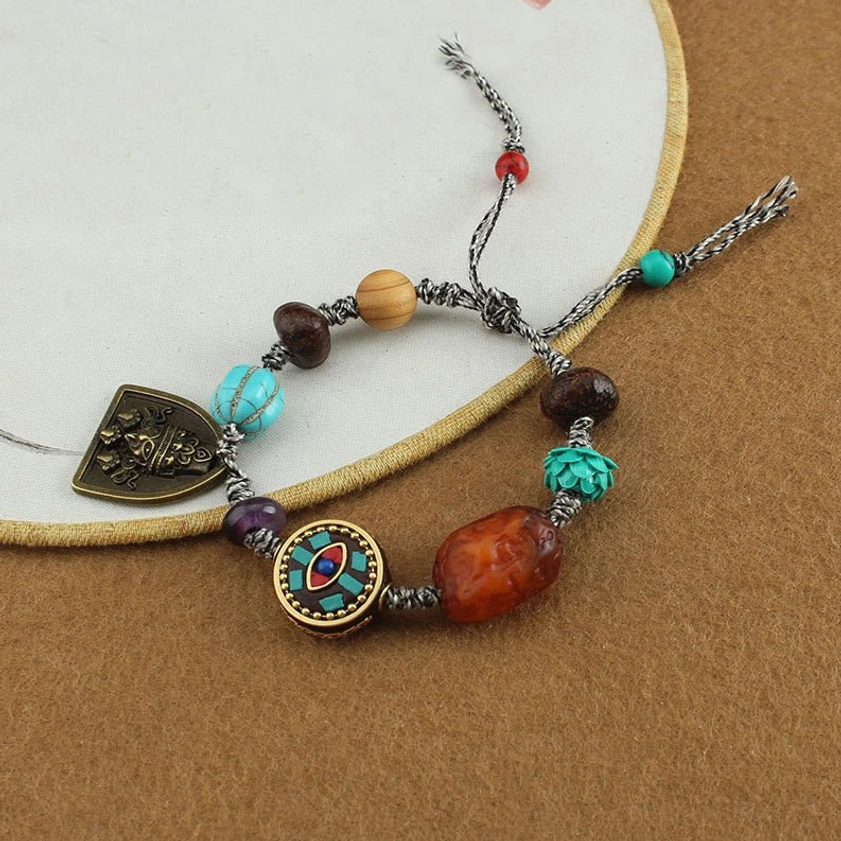 Buddhist protection rope bracelet worn angle view showing braided gray cord, enamel charm, mixed beads, and adjustable tassels
