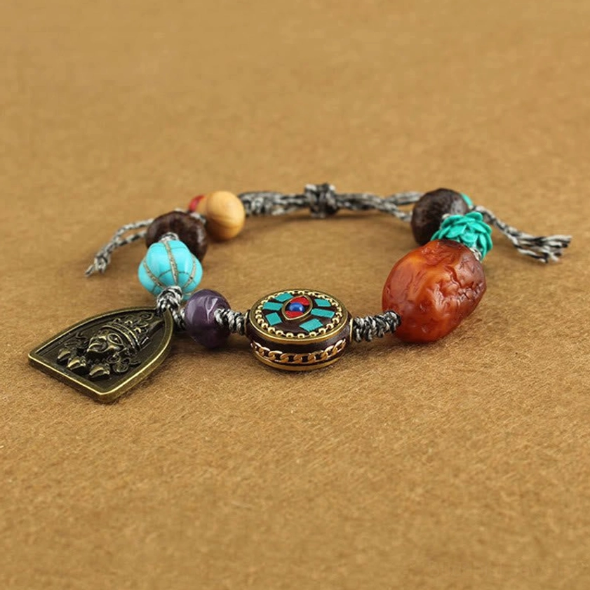 Oblique view of Buddhist rope bracelet featuring turquoise stone, rough amber bead, enamel round charm, and vintage metal deity pendant