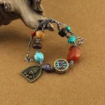 Buddhist protection bracelet with turquoise lotus bead and deity charm on brown fabric background, showing detailed braided rope and mixed gemstone beads