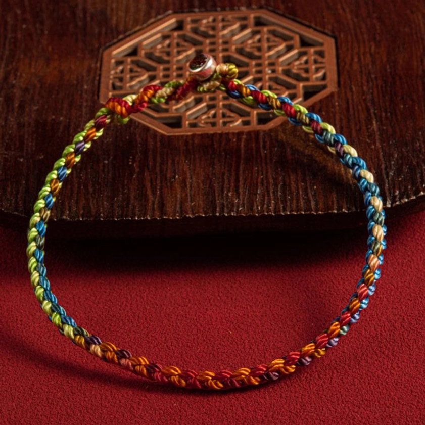 Handcrafted multicolored Buddhist bracelet with rainbow gradient braided rope and metal bead clasp on red fabric backdrop