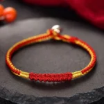 Handmade red and gold braided Buddhist protection bracelet with metal clasp, displayed on dark slate backdrop with spiritual fabric
