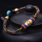 Handmade Buddhist red string bracelet with copper coin and eight knots, displayed on black teapot showing multicolor braided design with gold accents