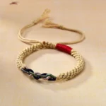 Hand-woven beige and red Buddhist cotton rope bracelet with twisted multi-color knot and adjustable tassel cords, displayed on tan fabric surface
