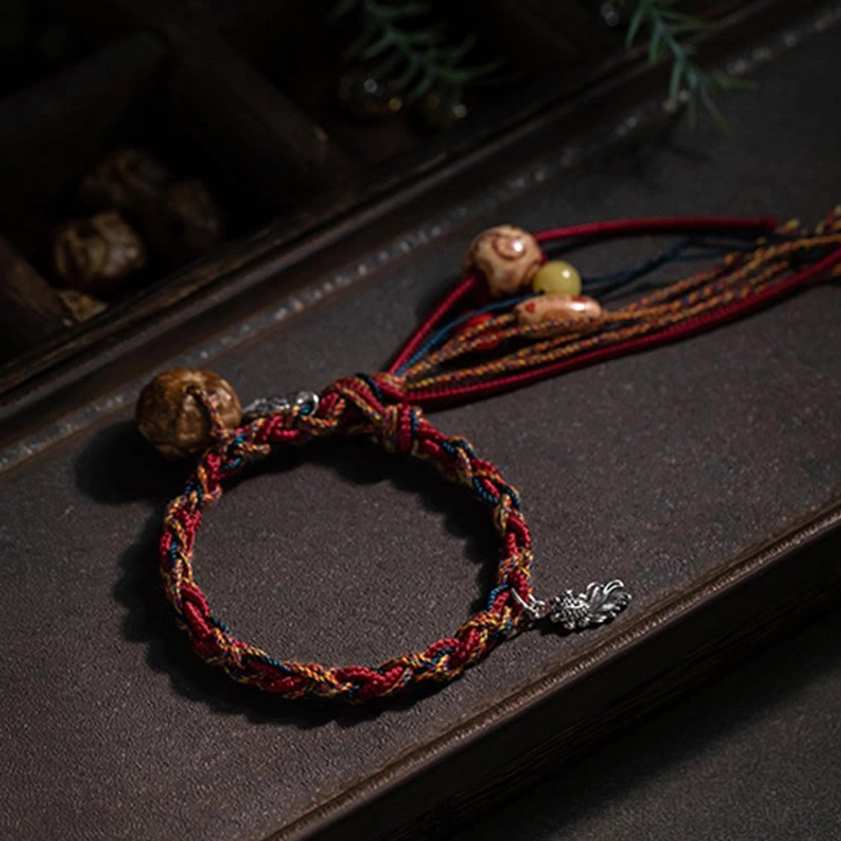 Buddhist red string bracelet with multicolor braided cords, silver leaf charm, amber-like beads and tassel, top-down styling on dark wooden background with foliage