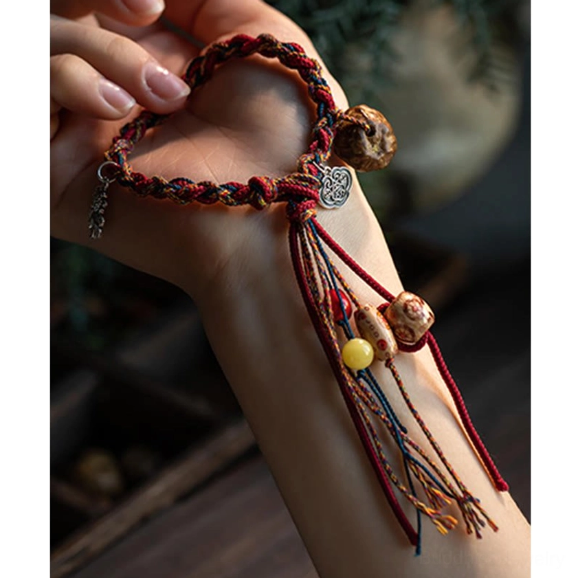 Red string Buddhist bracelet on wrist featuring silver Ruyi charm, yellow bead accent, carved wooden beads and decorative tassel, side arm position