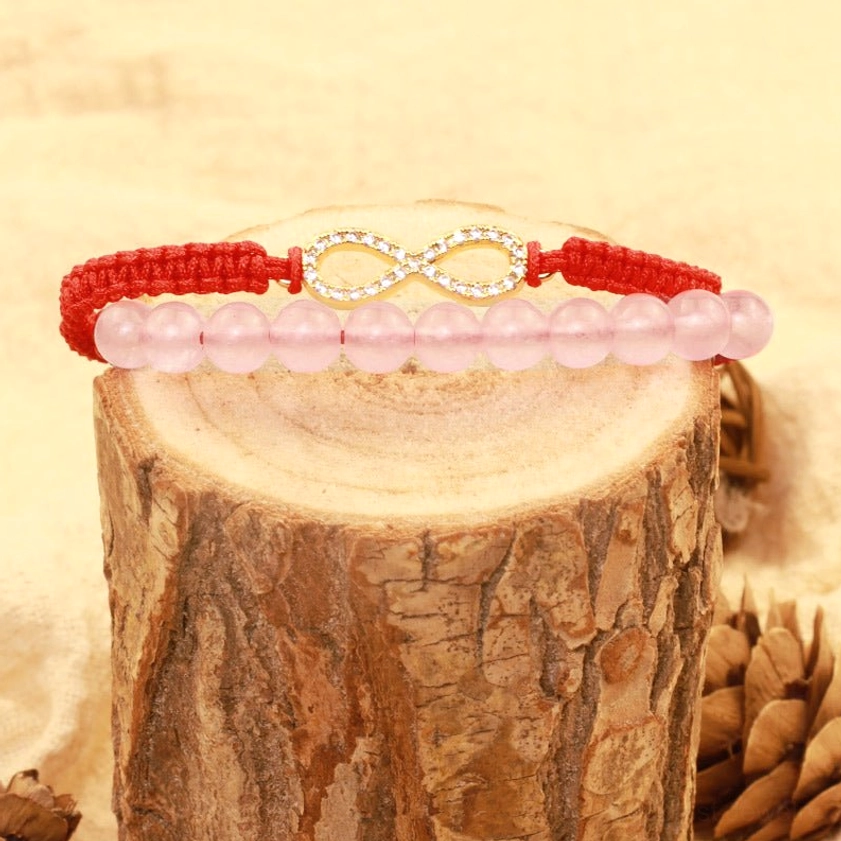Red string Buddhist bracelet worn at eye level showing gold infinity charm detail and smooth pink crystal beads on wooden backdrop