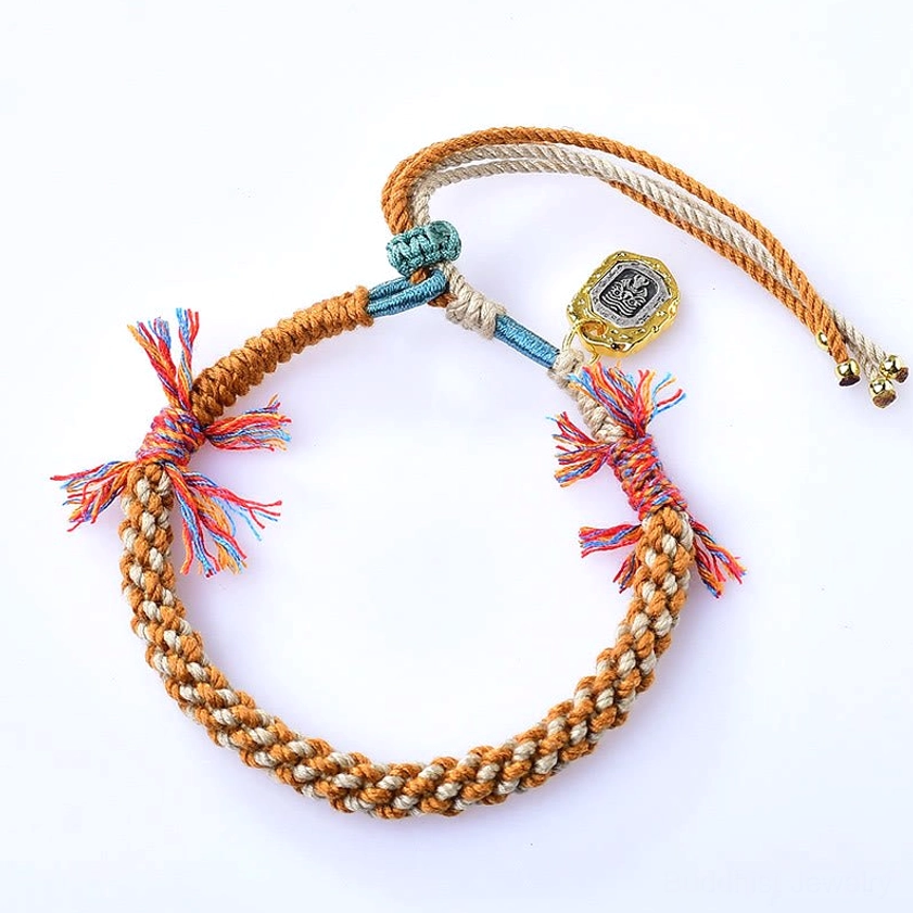 Handcrafted Buddhist string bracelet displaying visible engraved metal Zakiram charm, multicolor braided rope pattern, adjustable end straps with gold beads, and spiritual tassel decorations