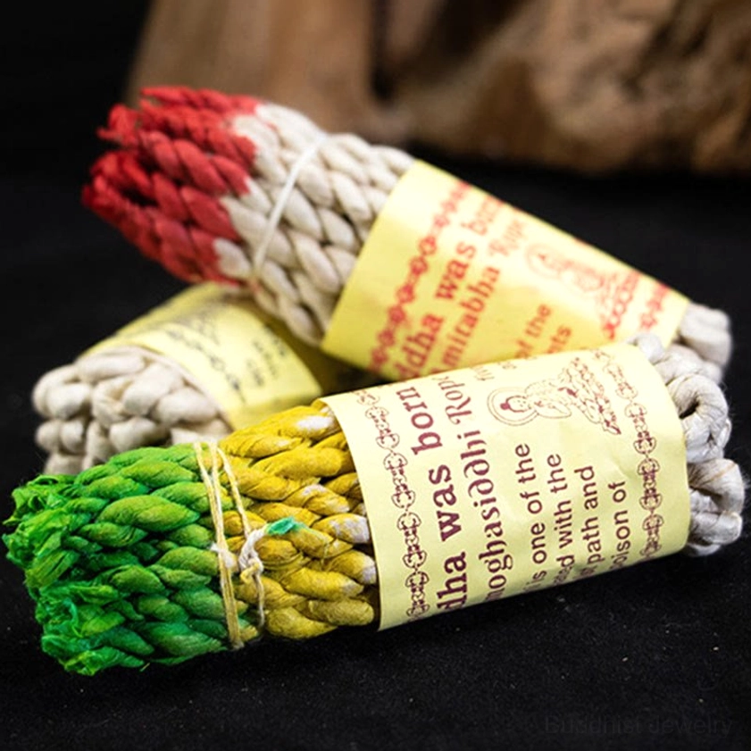 Multiple Buddhist rope incense bundles with colorful ends tied with natural twine on black background, showing handmade paper labels with Buddha illustrations