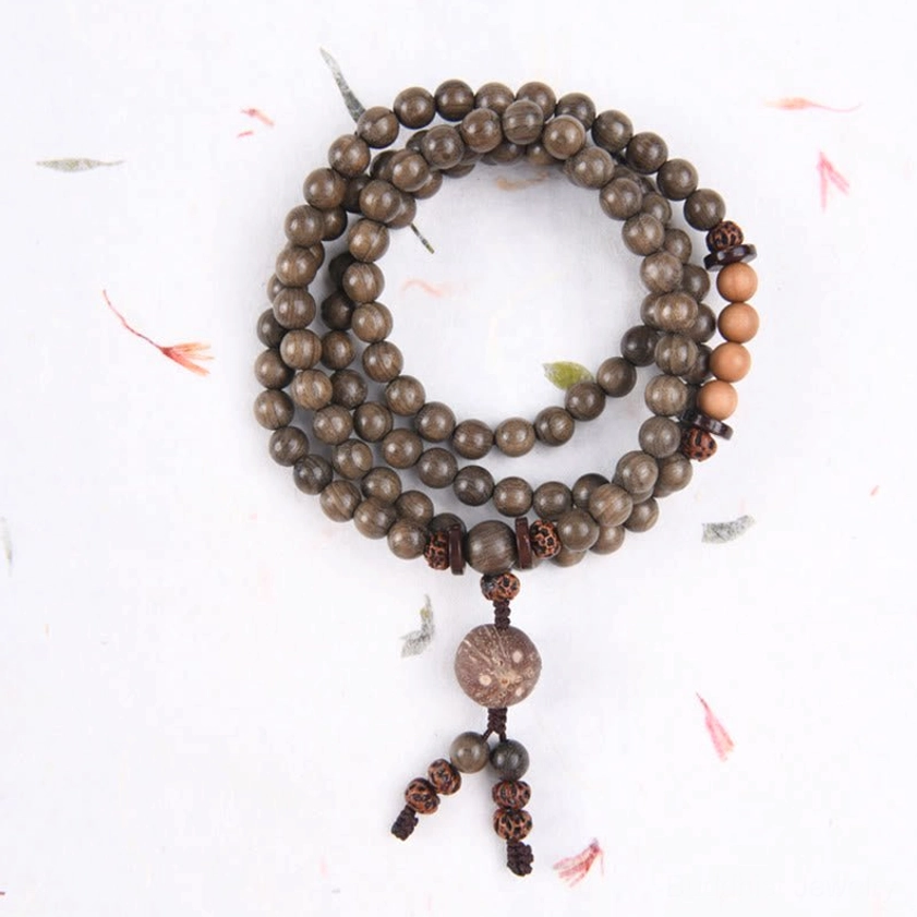 Single-loop sandalwood mala with small dark brown beads and patterned tassel pendant, overhead flat lay on white paper with flower petal decorations