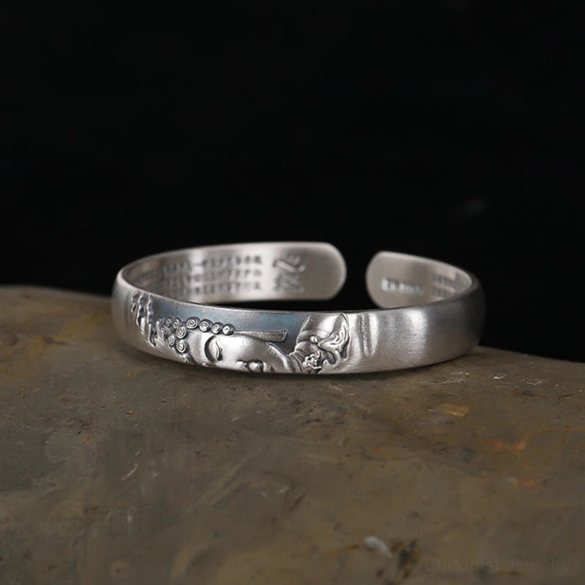 Top-down side view of open silver Buddha cuff bracelet, matte silver finish with embossed Buddha head and inner sutra engravings on stone background