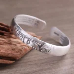 999 sterling silver Buddha head open cuff bracelet with heart sutra engravings, matte finish, left three-quarter view on wooden surface