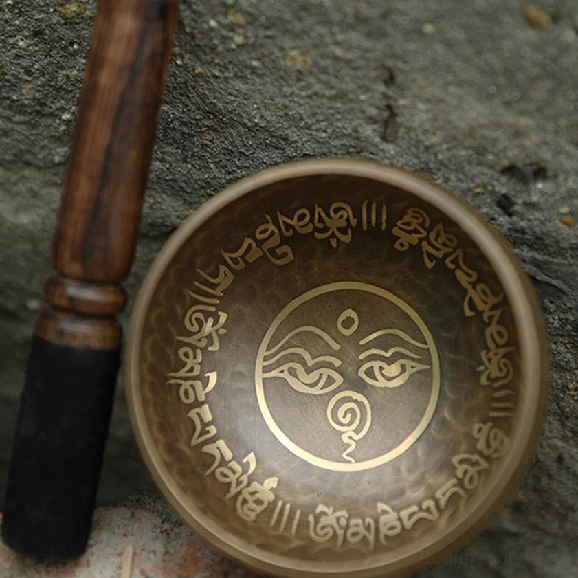 Handcrafted Buddhist singing bowl with wooden mallet striker on stone surface, showing hammered brass texture and golden sacred prints
